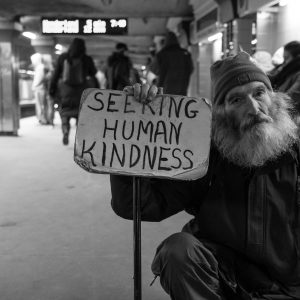 An unhoused man holds a sign that reads "Seeking human kindness" on the MBTA