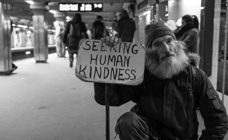 An unhoused man holds a sign that reads "Seeking human kindness" on the MBTA